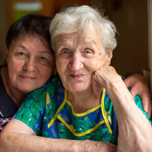 Photo of an elderly woman with white hair. Her adult daughter rests her head on her mother's shoulder.